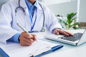 maos da medica irreconhecivel escrevendo no formulario e digitando no teclado do laptop (1)